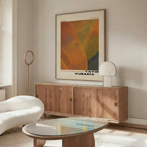 May include: Framed vintage-style poster with orange, yellow and teal dotted abstract design hanging above mid-century modern walnut credenza. Room scene shows white sculptural chair, round glass coffee table, floor lamp, and table lamp on wooden sideboard against beige wall.