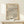 May include: Framed poster with flowing wavy lines in black, terracotta, and teal colors on cream background, displayed on light wood shelf against white wall. Small wrapped gift with evergreen sprig sits nearby. Natural wood frame complements minimalist modern interior styling.