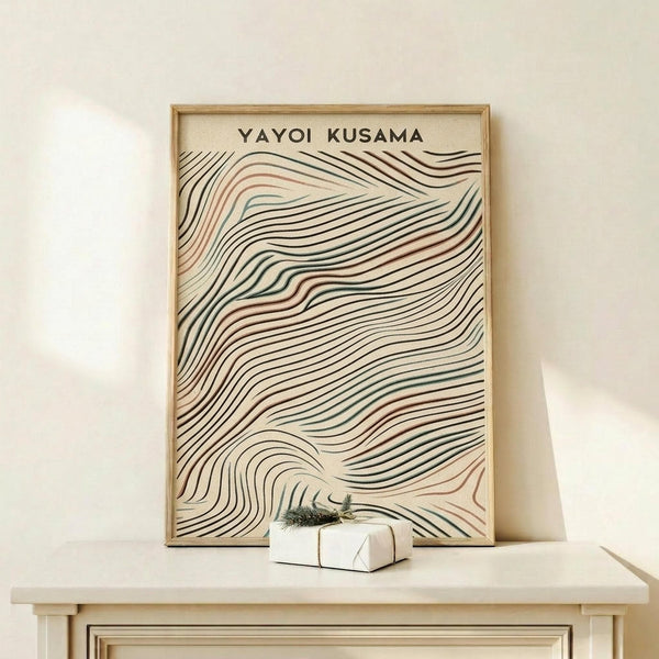 May include: Framed poster with flowing wavy lines in black, terracotta, and teal colors on cream background, displayed on light wood shelf against white wall. Small wrapped gift with evergreen sprig sits nearby. Natural wood frame complements minimalist modern interior styling.