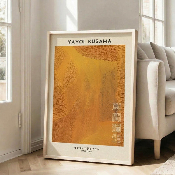 May include: Framed art print with orange dotted pattern artwork displayed on wooden floor against white wall next to beige upholstered sofa. Print features warm golden orange background with intricate dotted texture, Japanese text characters on right side, and title text at top and bottom. Light wood frame complements bright natural lighting from nearby windows.