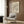 May include: Modern beige armchair with geometric patterned throw pillow positioned next to tall brass floor lamp with white marble base. Framed wall art featuring flowing black wavy lines on cream background hangs on wood-paneled wall. Dark round side table holds open book. Neutral curtains frame window with natural light illuminating warm wood floors and contemporary interior design setting.