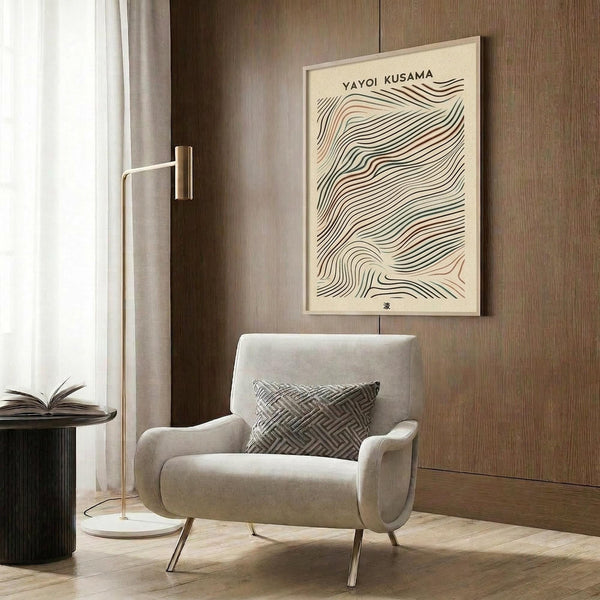 May include: Modern beige armchair with geometric patterned throw pillow positioned next to tall brass floor lamp with white marble base. Framed wall art featuring flowing black wavy lines on cream background hangs on wood-paneled wall. Dark round side table holds open book. Neutral curtains frame window with natural light illuminating warm wood floors and contemporary interior design setting.