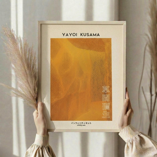 May include: Person holding framed art print with orange dotted pattern reminiscent of Kusama's infinity nets style, displayed against neutral curtained background with dried pampas grass. Light wooden frame contains cream matted artwork featuring flowing orange dots and Japanese text. Hands wearing beige ruffled sleeves show scale of medium-sized print in soft natural lighting.