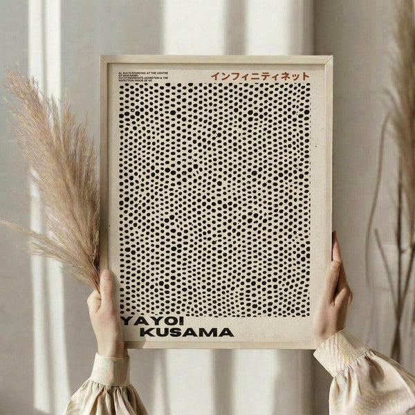 May include: Person in beige clothing with puffy sleeves holding a framed poster featuring dense black polka dots on cream background, with "YAYOI KUSAMA" text at bottom. Light wood frame, neutral interior setting with dried pampas grass visible to the left, soft natural lighting creating gentle shadows on white curtained background.