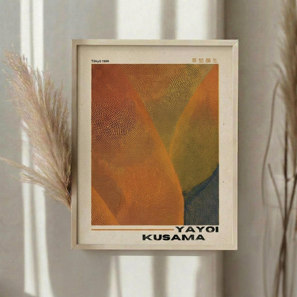 May include: Framed art print with abstract geometric design in warm orange, yellow and dark blue tones created with dotted halftone pattern. Print shows "YAYOI KUSAMA" text at bottom and "Tokyo 1998" at top. Displayed in light wood frame on white wall next to dried pampas grass stems, with soft natural lighting and neutral curtains in background.