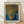 May include: Framed art print leaning against white wall on light wooden floor. The poster features abstract organic shapes in blue, orange and green with dotted texture patterns reminiscent of cellular structures. Text reads "Yayoi Kusama" at top and Japanese characters vertically on right side, with "infinity nets" at bottom. Natural wood frame with cream matting, soft natural lighting creates gentle shadows on wall.