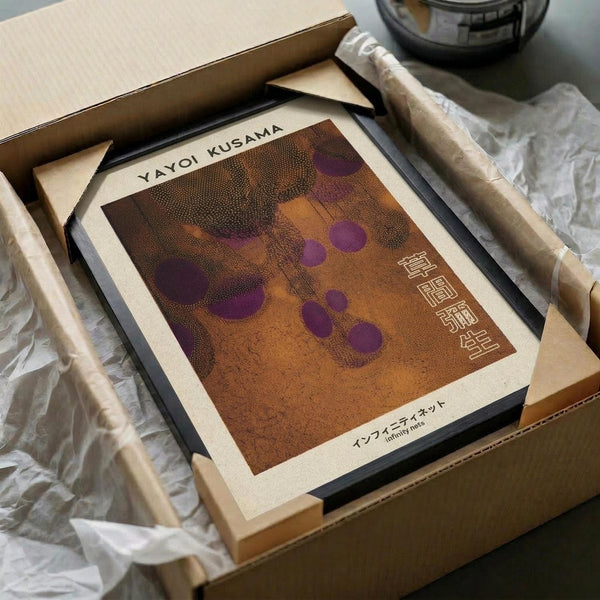 May include: Framed art print inspired by Yayoi Kusama displayed in wooden box with tissue paper packaging. Print features warm brown and orange tones with purple polka dots in dotted pattern style. Black frame sits among cardboard shipping materials and wooden dowels on neutral surface.