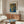 May include: Framed Yayoi Kusama exhibition poster with dotted abstract pattern in blue, orange, and green tones, displayed above a mid-century walnut credenza with sliding doors. White sculptural chair and glass-top coffee table complete the minimalist living room scene with cream walls and natural lighting.