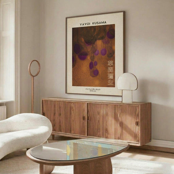 May include: Framed Yayoi Kusama poster featuring brown and orange tones with purple polka dots and Japanese text, displayed above a mid-century modern walnut credenza in a minimalist living room with white walls, cream chair, glass coffee table, and warm natural lighting