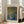 May include: Framed poster with abstract dotted pattern in blue, orange and yellow tones, featuring Japanese text and "Yayoi Kusama" at top. White wooden frame leans against cream wall next to beige upholstered sofa with pillows. Bright natural light streams through tall windows onto hardwood floors in modern living room setting.
