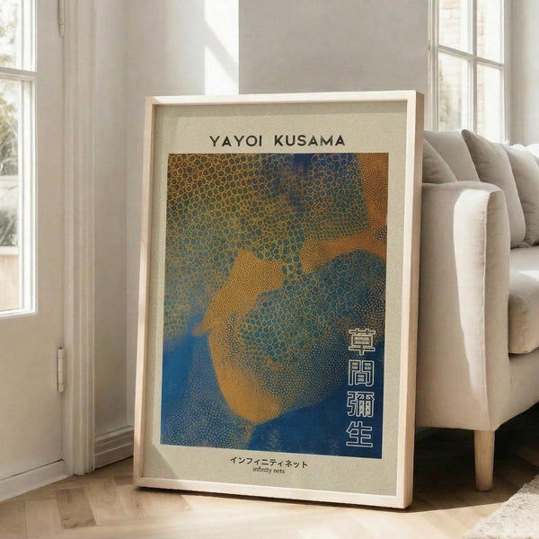 May include: Framed poster with abstract dotted pattern in blue, orange and yellow tones, featuring Japanese text and "Yayoi Kusama" at top. White wooden frame leans against cream wall next to beige upholstered sofa with pillows. Bright natural light streams through tall windows onto hardwood floors in modern living room setting.