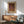 May include: Framed Yayoi Kusama exhibition poster featuring dotted sculptural forms in warm browns and purples, displayed above a mid-century wooden credenza with ceramic lamp. Green upholstered armchair with wooden frame sits nearby in sunlit modern living room with white walls and round coffee table.