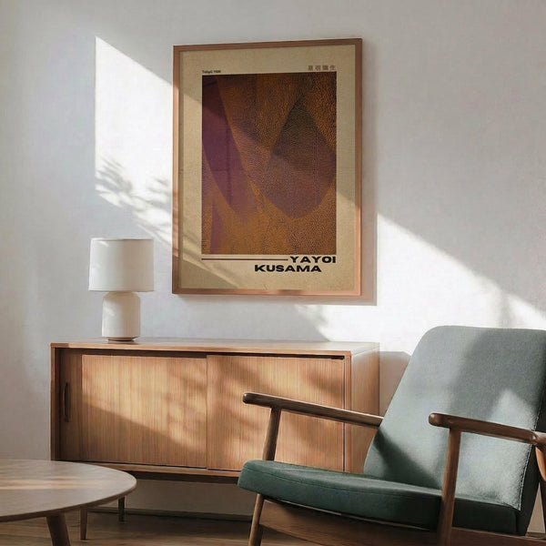 May include: Framed Yayoi Kusama exhibition poster featuring dotted sculptural forms in warm browns and purples, displayed above a mid-century wooden credenza with ceramic lamp. Green upholstered armchair with wooden frame sits nearby in sunlit modern living room with white walls and round coffee table.