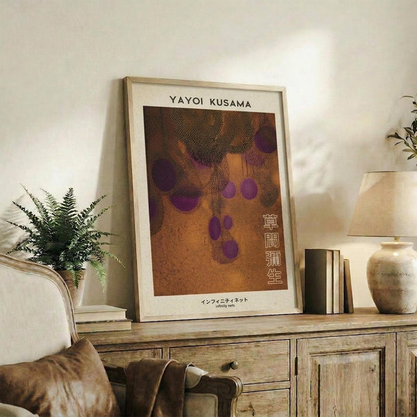 May include: Framed poster with warm brown and orange background featuring purple polka dot pattern, displayed on rustic wooden console table alongside fern plant, books, and ceramic vase in cozy living room setting with beige armchair visible in foreground.