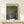 May include: Framed poster with "YAYOI KUSAMA" text at top and Japanese characters on right side, featuring abstract dotted pattern in blue, orange and gold tones resembling organic flowing shapes. Wooden frame sits on light wood surface next to small white wrapped gift with twine and greenery, against cream wall with natural lighting.