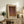 May include: Framed art print with abstract organic shapes in warm brown, purple and orange tones displayed on rustic wooden sideboard alongside potted fern, ceramic vases, and books in cozy living room setting with beige chair and neutral walls