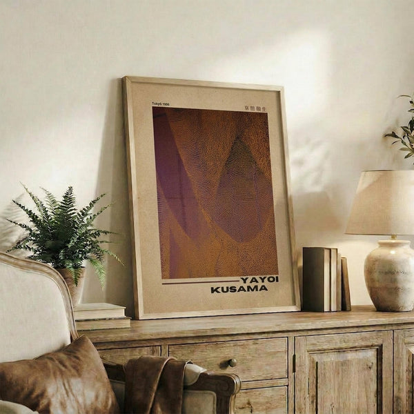 May include: Framed art print with abstract organic shapes in warm brown, purple and orange tones displayed on rustic wooden sideboard alongside potted fern, ceramic vases, and books in cozy living room setting with beige chair and neutral walls