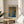 May include: Framed poster with Yayoi Kusama's name at top and colorful abstract dotted pattern in blues, oranges and yellows with Japanese text, displayed on round wooden side table next to beige upholstered chair with white pillow, plant leaves visible in corner, soft natural lighting creating shadows on white wall