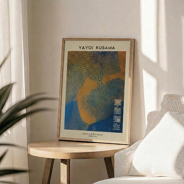 May include: Framed poster with Yayoi Kusama's name at top and colorful abstract dotted pattern in blues, oranges and yellows with Japanese text, displayed on round wooden side table next to beige upholstered chair with white pillow, plant leaves visible in corner, soft natural lighting creating shadows on white wall