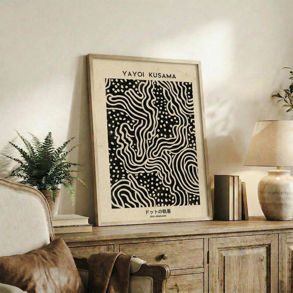 May include: Black and white abstract art print with flowing wavy lines and polka dots in wooden frame, displayed on rustic wood sideboard next to small fern plant, brown leather chair, ceramic vases and books in neutral-toned living room setting