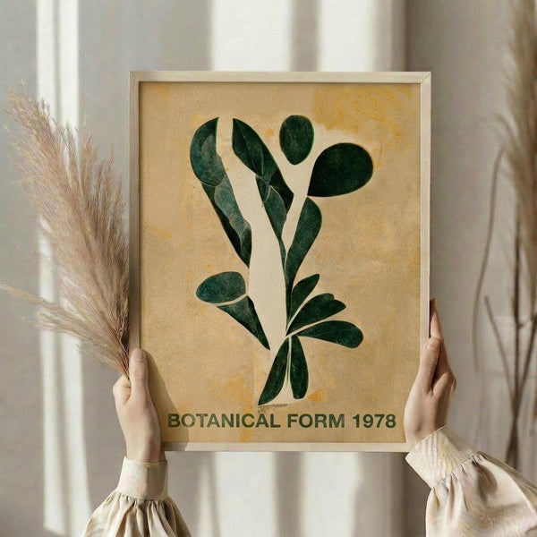 May include: Hands holding a framed botanical art print featuring dark green stylized leaf shapes arranged in an organic pattern against a warm beige textured background. The print is displayed in a simple light wood frame. Person wearing cream-colored ruffled sleeves holds dried wheat stalks in one hand while presenting the artwork near a bright window with soft natural lighting.