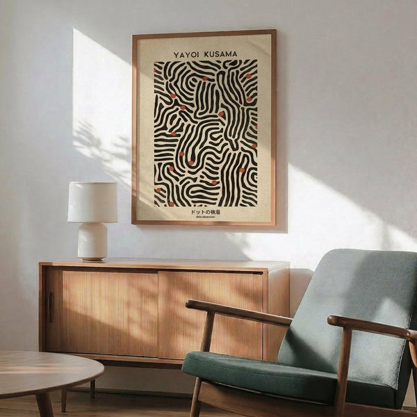 May include: Framed abstract art print featuring black curved lines with red accents on cream background, displayed above wooden credenza with white ceramic table lamp. Mid-century modern green upholstered armchair with wooden frame sits nearby on light wooden floors. Warm natural sunlight creates geometric shadows on white wall.