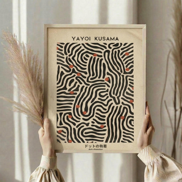 May include: Hands holding a framed art print featuring flowing black lines with scattered orange-red dots on cream background, displayed against white curtains with dried pampas grass visible to the left. The wooden frame appears natural finish, and Japanese text appears below the abstract pattern.