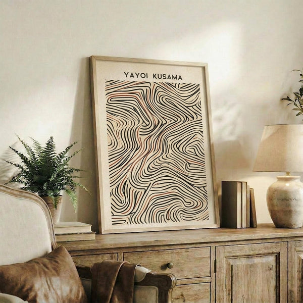 May include: Framed art print with flowing wavy lines in brown and black tones on cream background, displayed on rustic wooden sideboard alongside decorative pottery, books, and fern plant in neutral-toned living room setting with beige upholstered furniture.