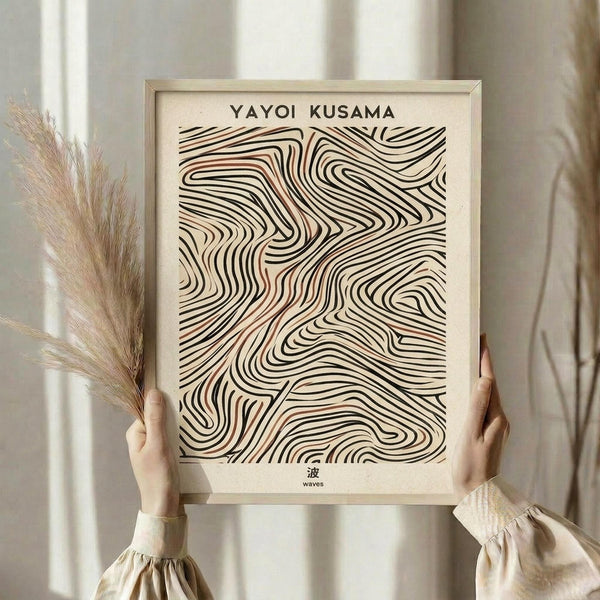 May include: Hands holding a framed abstract art print featuring flowing black and coral-colored wavy lines in an organic pattern. Light wood frame against neutral background with dried pampas grass visible. Person wearing cream-colored clothing with ruffled sleeves. Chinese character visible at bottom of print.