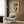 May include: Beige armchair with geometric pillow in modern living room setting with wood paneled walls. Framed botanical poster shows green leaf forms on warm yellow background with "BOTANICAL FORM 1978" text below. Gold floor lamp and dark round side table with open book visible. Natural lighting from window with white curtains creates warm, contemporary interior atmosphere.