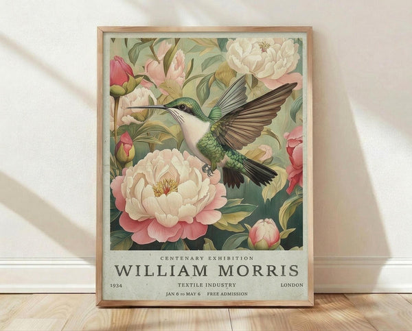 May include: Framed vintage-style exhibition poster featuring a green and white hummingbird with outstretched wings hovering among pink and white peonies with lush foliage. The botanical illustration has muted, naturalistic colors against a sage green background. Below the artwork, text reads "Centenary Exhibition William Morris" with exhibition details. The poster sits in a light wood frame on wooden flooring against a white textured wall with soft shadows.