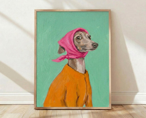 May include: Framed poster print leaning against white wall on wooden floor. Artwork shows Italian Greyhound dog wearing bright pink headscarf and orange sweater against mint green background. Painting has visible brushstrokes and portrait-style composition. Natural wood frame. Soft lighting creates gentle shadows on wall.