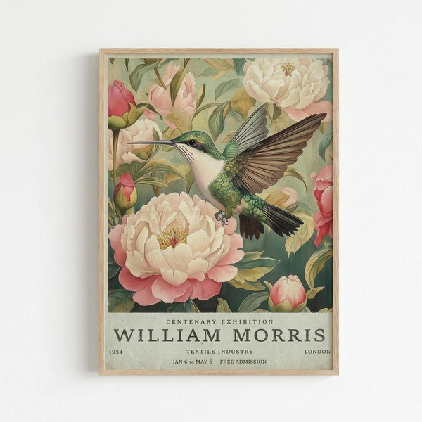 May include: Framed poster print featuring a detailed hummingbird with iridescent green and teal feathers hovering among large pink and cream peonies with lush green foliage. The vintage-style artwork has soft, muted colors on a sage green background. White text at bottom reads "CENTENARY EXHIBITION WILLIAM MORRIS" with exhibition details. Light wood frame hangs against white wall.