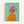 A framed painting of an Italian Greyhound wearing a bright pink headscarf and orange sweater against a mint green background. The dog has large expressive eyes and tan and brown coloring. The artwork is displayed in a simple wooden frame against a white wall, showing visible paint texture and brushstrokes in a portrait style.