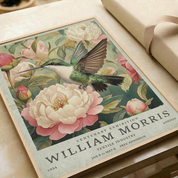 May include: Vintage-style poster print featuring a green and white hummingbird with outstretched wings hovering among large pink and white peonies with green foliage. Text reads "William Morris Centenary Exhibition London 1934" with exhibition details. Print shown on cream paper alongside neutral ribbon on light wooden surface.