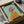 May include: Framed artwork of an Italian Greyhound wearing a bright pink headscarf and orange sweater against a mint green background, shown being carefully unpacked from a cardboard shipping box with protective tissue paper and wooden frame corners visible.