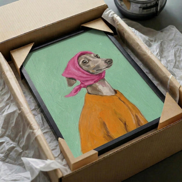 May include: Framed artwork of an Italian Greyhound wearing a bright pink headscarf and orange sweater against a mint green background, shown being carefully unpacked from a cardboard shipping box with protective tissue paper and wooden frame corners visible.