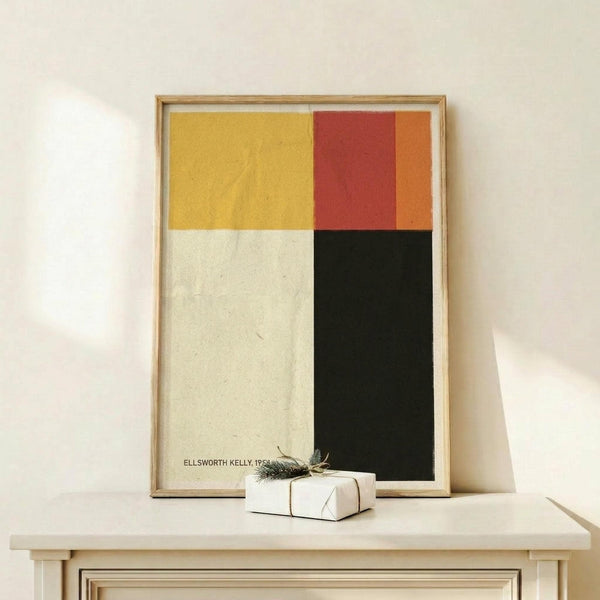 May include: Framed abstract art print with four colored blocks - yellow, red-orange, cream, and dark green - displayed on light wood surface against white wall. Small wrapped gift with evergreen sprig sits nearby. Natural wood frame with simple lines complements minimalist geometric design.