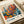 May include: Vintage-style exhibition poster with abstract botanical design featuring colorful circular flowers and foliage in orange, blue, teal, and yellow tones. Text shows Japanese characters and "MUSÉE D'ART MODERNE DE LA VILLE DE PARIS" with "JAPAN 1984" and date "5. JUIN - 19. JUIN 1986". Poster appears framed in light wood, displayed on white surface with rolled paper tubes nearby.