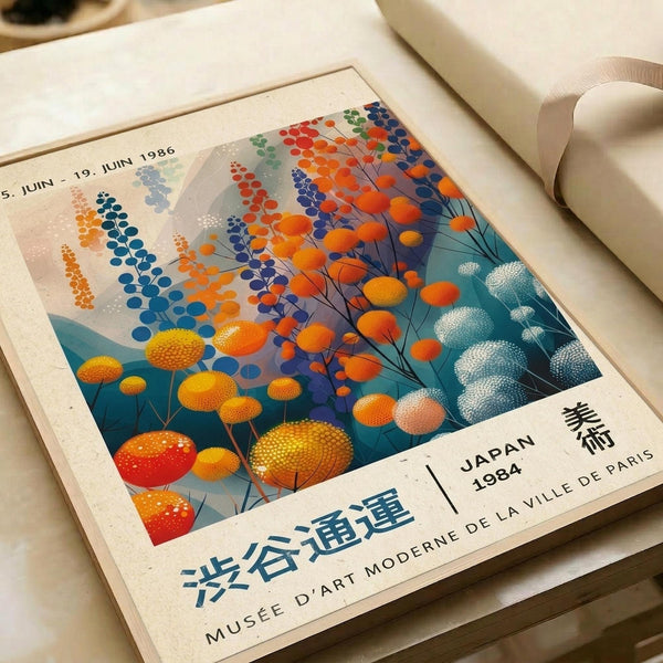 May include: Vintage-style exhibition poster with abstract botanical design featuring colorful circular flowers and foliage in orange, blue, teal, and yellow tones. Text shows Japanese characters and "MUSÉE D'ART MODERNE DE LA VILLE DE PARIS" with "JAPAN 1984" and date "5. JUIN - 19. JUIN 1986". Poster appears framed in light wood, displayed on white surface with rolled paper tubes nearby.