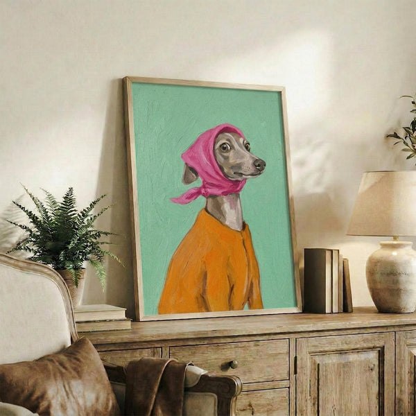May include: Framed poster print displaying an Italian Greyhound wearing a bright pink headscarf and orange sweater against a mint green background, styled as a portrait painting. The artwork sits on a rustic wooden console table alongside books, ceramic vases, and a fern plant, with a cream upholstered chair visible in a cozy living room setting with white walls.
