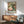 May include: Vintage-style poster featuring a green hummingbird with spread wings hovering near pink and white peonies with green foliage, displayed in wooden frame above mid-century modern credenza with white ceramic lamp, next to sage green upholstered armchair with wooden arms in bright, naturally-lit room with white walls.