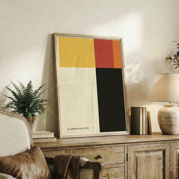 May include: Framed abstract art print with yellow, red, orange, white and black color blocks displayed on rustic wooden sideboard alongside fern plant, books, ceramic vases and brown leather chair in warm, naturally-lit living room setting