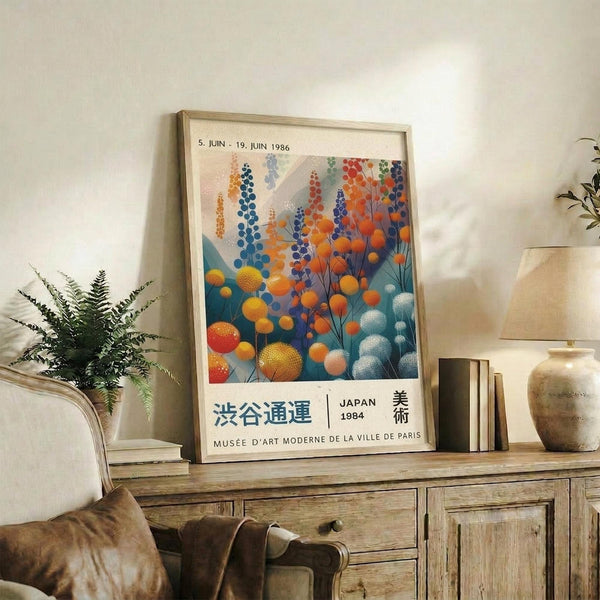 May include: Framed vintage-style Japanese art poster featuring colorful botanical illustration with orange, blue, and teal spherical flowers and foliage, displayed on rustic wooden console table alongside fern plant, ceramic vase, books, and beige upholstered chair in neutral-toned living room setting
