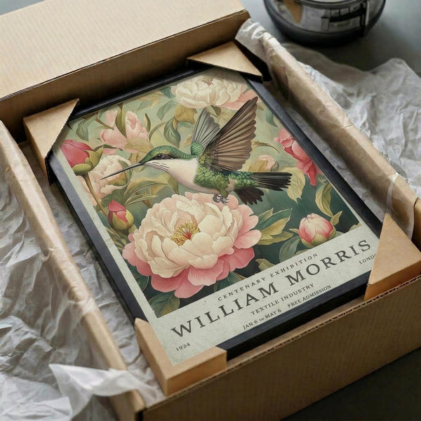 May include: Vintage-style botanical poster featuring a green hummingbird with spread wings hovering among pink and cream peonies with green foliage. The artwork has "William Morris" text at bottom and is displayed in a black frame nestled in an open cardboard shipping box with white tissue paper padding.