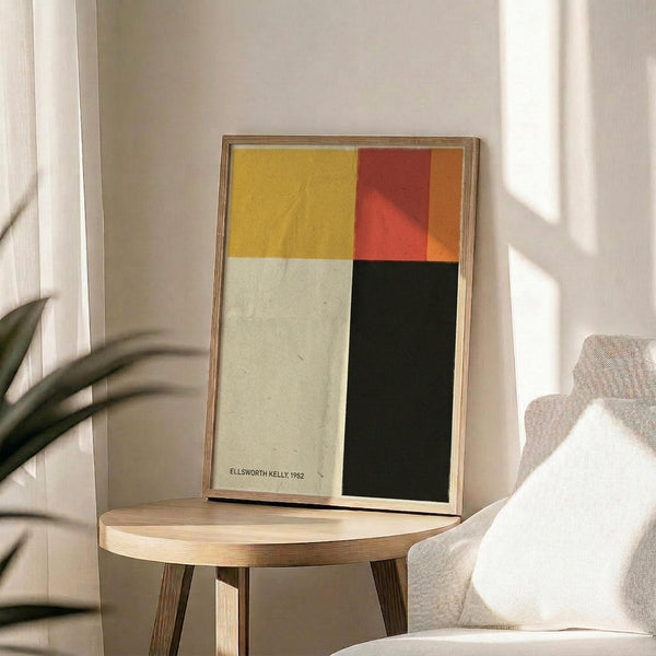 May include: Abstract color block artwork in wooden frame displayed on round wooden side table next to beige upholstered chair. Poster features four geometric sections in mustard yellow, red-orange gradient, cream white, and dark forest green. Natural lighting creates soft shadows across neutral-toned room with plant leaves visible in corner.