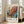 May include: Large framed poster featuring abstract botanical artwork with colorful spherical flowers in orange, blue, and teal tones against a soft background. Shows exhibition text "5. JUIN - 19. JUIN 1986" at top, Japanese characters, "JAPAN 1984," and "MUSÉE D'ART MODERNE DE LA VILLE DE PARIS" at bottom. Light wood frame leans against white wall next to beige upholstered sofa in bright, naturally-lit room with windows.