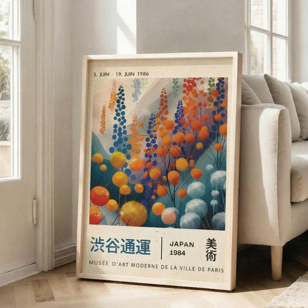 May include: Large framed poster featuring abstract botanical artwork with colorful spherical flowers in orange, blue, and teal tones against a soft background. Shows exhibition text "5. JUIN - 19. JUIN 1986" at top, Japanese characters, "JAPAN 1984," and "MUSÉE D'ART MODERNE DE LA VILLE DE PARIS" at bottom. Light wood frame leans against white wall next to beige upholstered sofa in bright, naturally-lit room with windows.