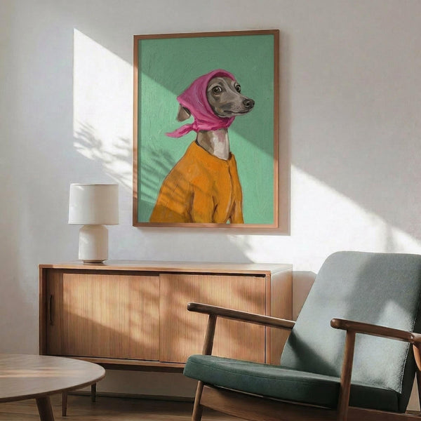 May include: Framed artwork showing an Italian Greyhound wearing a pink headscarf and orange sweater against a mint green background, displayed above a mid-century modern wood credenza with table lamp, next to a gray upholstered armchair with wooden frame in a sunlit room with white walls.