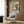 May include: Framed botanical poster featuring a green hummingbird with spread wings among pink and white peonies, displayed above a beige upholstered armchair with geometric pillow in a modern living room with wood paneling, brass floor lamp, and side table with open book.